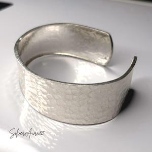 May include: A wide, open-ended silver cuff bracelet featuring a hammered, textured surface across the exterior. The metal has a polished, reflective finish. The text "SilverAura925" is printed in a cursive script in the bottom left corner.