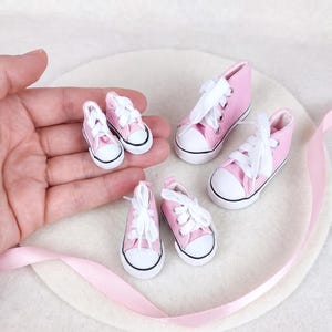 May include: A collection of miniature high-top trainers in light pink canvas with white rubber toe caps, white soles, and white laces. The set includes three pairs of shoes in varying small sizes, featuring silver-toned metal eyelets for the laces.