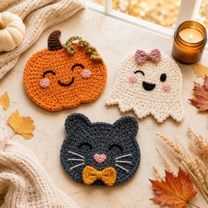 May include: Three crocheted coasters featuring seasonal characters: an orange pumpkin with a brown stem, a cream-colored ghost with a pink bow, and a dark gray cat with a yellow bow tie. Each piece has embroidered facial features and pink cheeks.