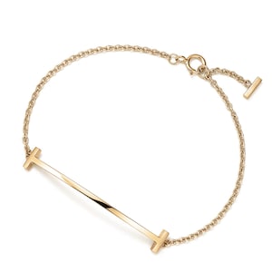 May include: A gold-toned chain bracelet featuring a horizontal bar element with T-shaped ends. The delicate link chain connects to the central bar and includes a circular spring ring clasp with a small dangling T-shaped charm at the closure.