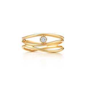 May include: A gold ring featuring a multi-band design with three flowing, interconnected gold wires. A single, round, brilliant-cut clear gemstone is bezel-set in the centre, positioned between the top and middle gold bands of the ring.