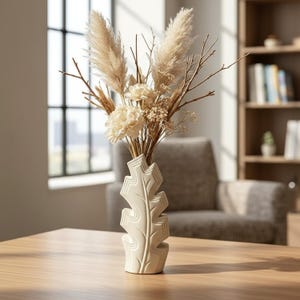 Op de afbeelding: Een crèmekleurige keramische vaas met een gestileerd bladvormig silhouet en verticale ribbeldetails. De vaas bevat een droogbloemenarrangement bestaande uit beige pampasgras, dunne bruine takjes en kleine gebroken witte bloesems.