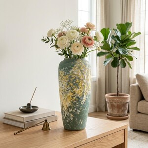 May include: A tall cylindrical vase with a textured matte finish in shades of sage green, white, and yellow, filled with a bouquet of white and pale pink ranunculus flowers and baby's breath, standing on a light wood surface.