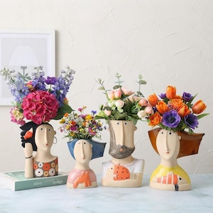 Op de afbeelding: Vier keramische vazen in de vorm van een hoofd met gestileerde gelaatstrekken en kleurrijke kledingpatronen. Elke vaas bevat een apart bloemstuk, waaronder roze hortensia's, kleine wilde bloemen, zachte rozen en levendige oranje tulpen.