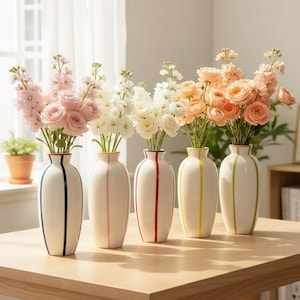 Könnte beinhalten: Fünf weiße Keramikvasen mit schlanker Silhouette, jede mit einem einzelnen vertikalen Streifen in Marineblau, Rosa, Rot, Gelb oder Grün. Jede Vase enthält einen Blumenstrauß in passenden Farben, darunter rosa, weiße und pfirsichfarbene Rosen.