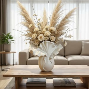 Może przedstawiać: Biały ceramiczny wazon z falistym, przypominającym muszlę brzegiem, wypełniony dużą kompozycją z suszonej beżowej trawy pampasowej, małych białych kwiatów i delikatnych łodyg. Wazon ma gładkie, matowe wykończenie oraz szeroki, zakrzywiony otwór.