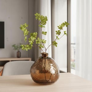 May include: A round, amber-colored glass vase with a textured, dimpled surface and a narrow neck. The vase contains a single slender branch with multiple small, bright green leaves extending upwards from the opening of the glass vessel.