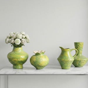 May include: A collection of four lime green and yellow ceramic vessels with a textured, dimpled surface. One wide vase holds a bouquet of white ranunculus flowers, while the others include a smaller scalloped vase, a pitcher, and a tall vase.
