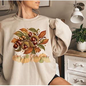 Könnte beinhalten: Ein beigefarbenes Sweatshirt mit Rundhalsausschnitt und einem zentralen Aufdruck aus braunen Kastanien, stacheligen Hüllen und herbstlichen Blättern in Orange-, Rot- und Grüntönen. Unter dem Motiv steht der Schriftzug "cosy season" in Schreibschrift über Kürbissen.