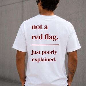 Könnte beinhalten: Ein weißes kurzärmeliges T-Shirt mit dunkelrotem Text auf der Rückseite, der lautet: not a red flag. gefolgt von einer horizontalen Linie und dem Text: just poorly explained. Das T-Shirt hat einen Rundhalsausschnitt und eine Standardpassform.