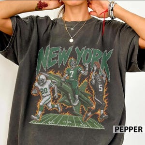 May include: A charcoal gray short-sleeve t-shirt featuring a graphic print of three skeleton football players in green uniforms with numbers 20, 7, and 5. The design includes a fighter jet, orange flames, a football field, and the text NEW YORK.