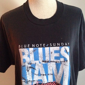 May include: Black t-shirt with a blue and white graphic print that says "Blues Jam" with the text "The Derelicts Band" underneath. The text "Blue Note Sunday" is at the top of the graphic.