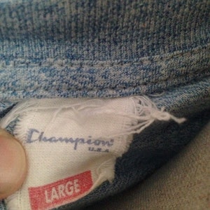 Pu&ograve; includere: Un'etichetta bianca del marchio Champion con testo blu e un'etichetta rossa che dice "LARGE" sotto. L'etichetta &egrave; attaccata a una camicia di jeans blu.