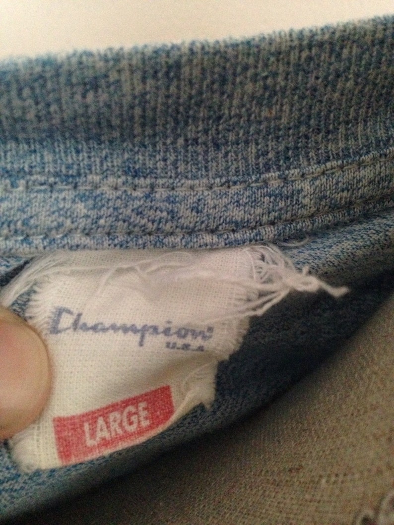 Pu&ograve; includere: Un'etichetta bianca del marchio Champion con testo blu e un'etichetta rossa che dice "LARGE" sotto. L'etichetta &egrave; attaccata a una camicia di jeans blu.