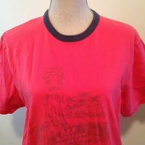 May include: A red t-shirt with a dark blue crew neck and short sleeves. The shirt features a faded graphic of a tiki statue and the text "A&F Bungalow Getaways". The shirt is displayed on a mannequin.