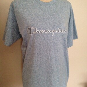 Pu&ograve; includere: Una maglietta blu chiaro con un logo Champion bianco.