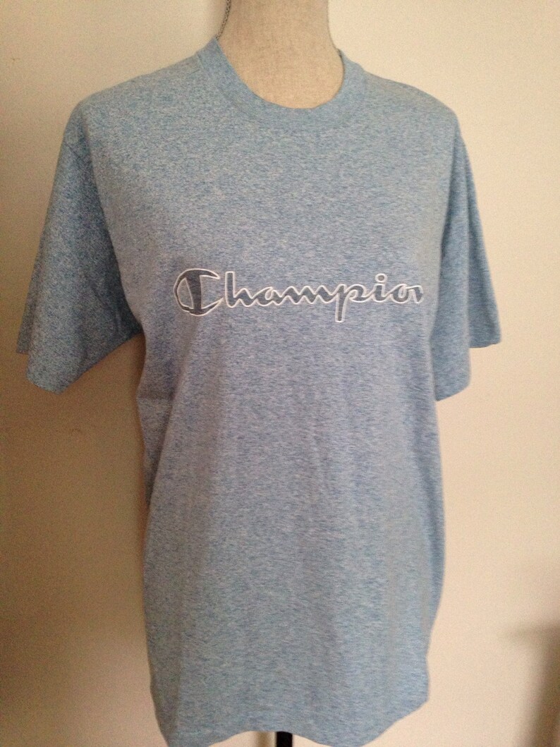Pu&ograve; includere: Una maglietta blu chiaro con un logo Champion bianco.