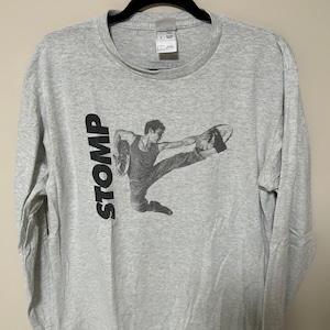 May include: Grey long-sleeved t-shirt with a black and white image of a person performing a dance move. The word "STOMP" is printed vertically on the left side of the shirt.