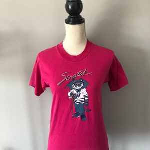 May include: A pink t-shirt featuring a cartoon cat wearing a hockey jersey and holding a hockey stick. The text "Scratch" is above the cat.