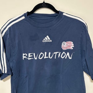 May include: A navy blue t-shirt with white stripes on the sleeves and a white adidas logo on the front. The shirt also has a white graphic of a football and an American flag with the word "REVOLUTION" written underneath.