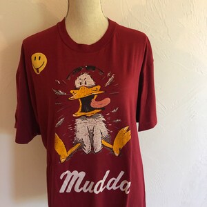 May include: A maroon t-shirt with a cartoon duck wearing sunglasses and a yellow smiley face on the front. The text "Mudda" is printed in white below the duck.