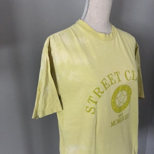 May include: A yellow t-shirt with a green graphic that says "Street Club" and features a star inside a circle with the text "There is the Real" and "Since MCMLVIII".