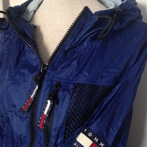 May include: A blue nylon jacket with a black mesh panel and a Tommy Hilfiger logo on the sleeve. The jacket has a zipper closure and a hood.