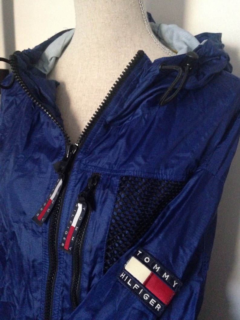 May include: A blue nylon jacket with a black mesh panel and a Tommy Hilfiger logo on the sleeve. The jacket has a zipper closure and a hood.