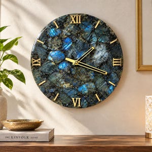 Puede incluir: Un reloj de pared redondo con una esfera de piedra labradorita texturizada con inclusiones iridiscentes azules y doradas. Presenta números romanos dorados en las posiciones de las doce, tres, seis y nueve, con manecillas doradas finas.