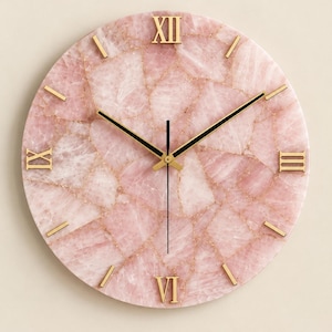 May include: A round wall clock featuring a face made of mosaic rose quartz pieces in shades of pale pink. The clock has gold-colored Roman numerals at the twelve, three, six, and nine o'clock positions, with gold-colored hour markers and thin black hands.