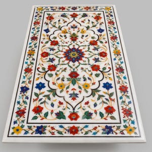 Könnte beinhalten: Eine rechteckige Tischplatte aus weißem Marmor mit einem komplexen floralen Einlegemuster. Die Oberfläche zeigt ein zentrales sternförmiges Motiv, umgeben von symmetrischen Ranken, Blättern und Blüten in leuchtenden Rot-, Blau-, Gelb- und Grüntönen.