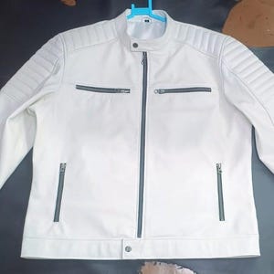 Könnte beinhalten: Eine weiße Jacke mit Stehkragen und Druckknopfverschluss, durchgehendem Frontreißverschluss und zwei Reißverschlusstaschen auf der Brust. Die Schultern und Oberärmel sind gesteppt, zudem gibt es zwei weitere Reißverschlusstaschen unten.