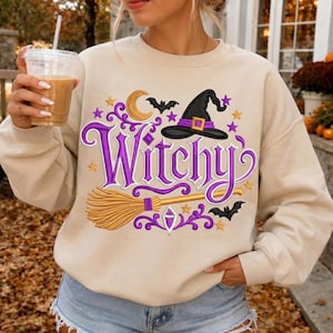 Op de afbeelding: Een beige sweater met ronde hals en een grafisch ontwerp met de paarse tekst Witchy, een zwarte heksenhoed, een beige bezemsteel, een gele maansikkel, kleine gouden sterren en zwarte vleermuizen, aangevuld met paarse decoratieve krulaccenten.