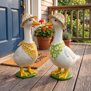 May include: Two white duck figurines standing on green bases. Each duck wears a straw hat adorned with white daisy flowers and a chin strap. One duck wears a yellow bandana with white daisies, while the other wears a green bandana with white daisies.