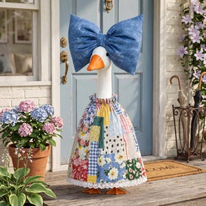 Puede incluir: Una figura de ganso blanco que lleva un lazo grande de textura vaquera azul en la cabeza y un vestido de retazos con varios patrones florales, de cuadros y de rayas en varios colores, acabado con un borde de encaje blanco en el dobladillo.