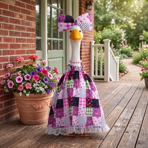 Op de afbeelding: Een wit ganzenbeeldje met een patchworkjurk met roze, groene en zwarte patronen, een bijpassende oversized strik op het hoofd en een kralenketting. De jurk heeft een geplooide witte kanten zoom en een geplooide halslijn.