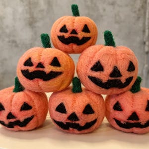 Könnte beinhalten: Ein Stapel aus sechs nadelgefilzten orangefarbenen Kürbissen, die in einer Pyramide angeordnet sind. Jeder Kürbis hat einen dunkelgrünen Filzstiel und ein schwarzes Filzgesicht mit dreieckigen Augen und einem breiten, gezackten Mund.