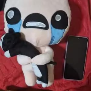 May include: A plush toy featuring a pale, round-headed character with large black eyes and blue tear-shaped markings, holding a smaller black and white cat-like plush toy, positioned next to a rectangular smartphone with a black screen.