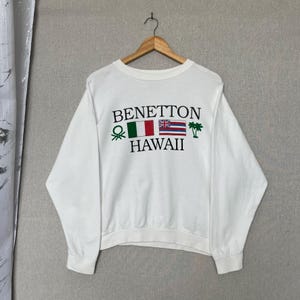 Puede incluir: Una sudadera blanca de manga larga con cuello redondo que presenta el texto BENETTON HAWAII en letras negras. El diseño incluye un logotipo verde, la bandera italiana, la bandera de Hawái y dos iconos de palmeras verdes en el pecho.