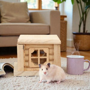 May include: A small, light-coloured hamster stands on a textured rug next to a miniature wooden log cabin house with a square window frame. To the right of the wooden structure sits a light purple, vertically ribbed ceramic mug with steam rising.