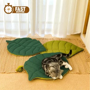 Puede incluir: Tres cojines de suelo con forma de hoja en tonos verde oscuro y verde claro, con patrones de nervaduras acolchadas. Un gato atigrado gris y negro descansa sobre uno de los cojines verde oscuro. Un icono marrón con el texto FAST SHIPPING.