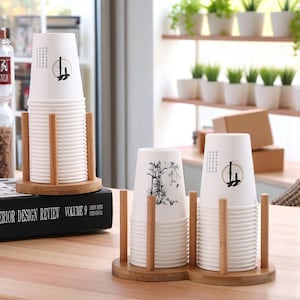 May include: Two light brown wooden cup holders with vertical dowels holding stacks of white paper cups. The cups feature black ink illustrations of bamboo stalks and Chinese characters. One holder is circular, and the other is an elongated oval shape.