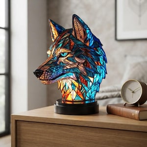 May include: A stained glass lamp shaped like a wolf head, featuring a geometric pattern of blue, orange, and yellow glass pieces. The wolf head is mounted on a circular black base, with bright blue eyes and a detailed snout, creating a vibrant display.