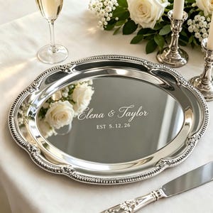 May include: A polished silver oval serving tray with a decorative beaded rim. The centre of the tray features the engraved text Elena & Taylor and EST 5. 12. 26. The reflective surface shows a clear reflection of white roses and greenery.