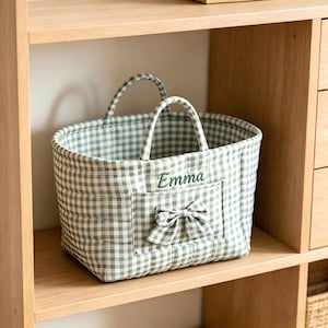 May include: A quilted fabric storage basket featuring a sage green and white gingham pattern. The basket has two handles, a front pocket with a matching gingham bow, and the name Emma embroidered in dark green thread on the front panel.