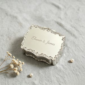 Puede incluir: Una caja rectangular de metal plateado con acabado pulido y un borde ornamentado con volutas en relieve. La tapa está grabada con el texto "Eleanor & James" en una fuente cursiva. La caja cuenta con una tapa con bisagras y una superficie reflectante.