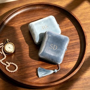 May include: Two rectangular light blue velvet jewellery boxes with gold embroidered initials BK and SD, accompanied by a gold pocket watch with a white face and a delicate pearl necklace, all arranged on a circular dark brown wooden tray.