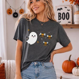 May include: A dark gray short-sleeve t-shirt featuring a graphic print of a smiling white ghost and three small yellow and black bees. The design includes small orange and white star accents scattered around the ghost and the bees.