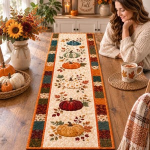 May include: A rectangular fabric table runner featuring a central cream-coloured panel decorated with a vertical row of six colourful pumpkins and autumn leaves. The runner is bordered by a patchwork design of small squares in shades of orange, green, and brown.