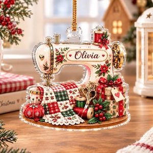 Può includere: Una decorazione natalizia piatta in acrilico a forma di macchina da cucire vintage, con il nome Olivia in corsivo. È decorata con stelle di Natale, un tessuto trapuntato rosso e verde, un puntaspilli, rocchetti di filo, forbici e pacchetti.